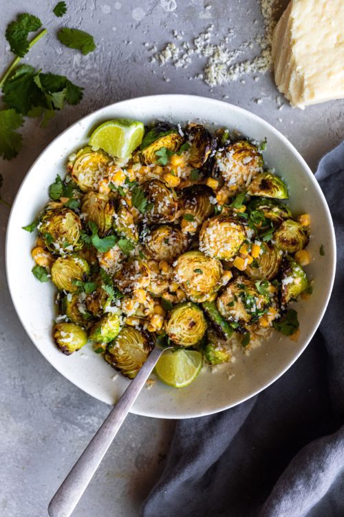 Mexican Street Corn Brussels Sprouts in a white bowl on a grey background and and wedges of fresh lime.