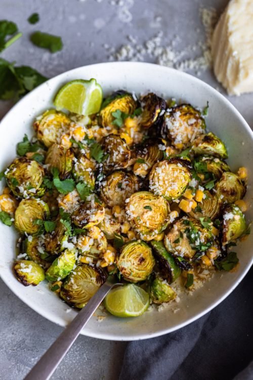 White bowl of Mexican Street Corn Brussels sprouts on a grey background with a metal spoon for serving.