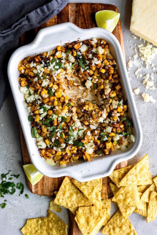 Hot Street Corn Dip in a white baking dish pictured on a wooden board and a grey background scattered with lime wedges, crumbled cheese and corn tortilla chips.