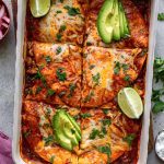 Overhead shot of an enchilada casserole garnished with sliced avocado and lime wedges.