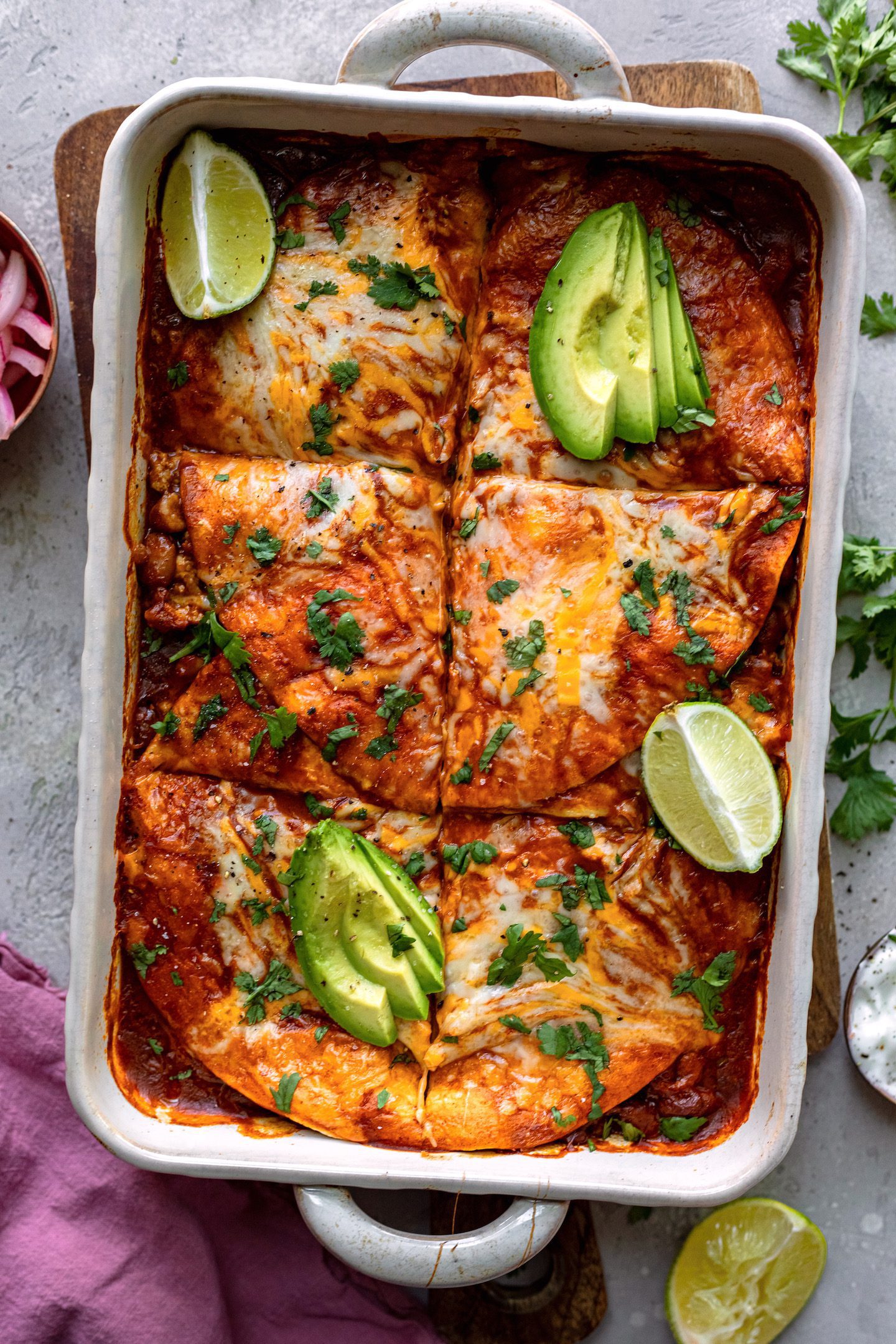 Overhead shot of an enchilada casserole garnished with sliced avocado and lime wedges.