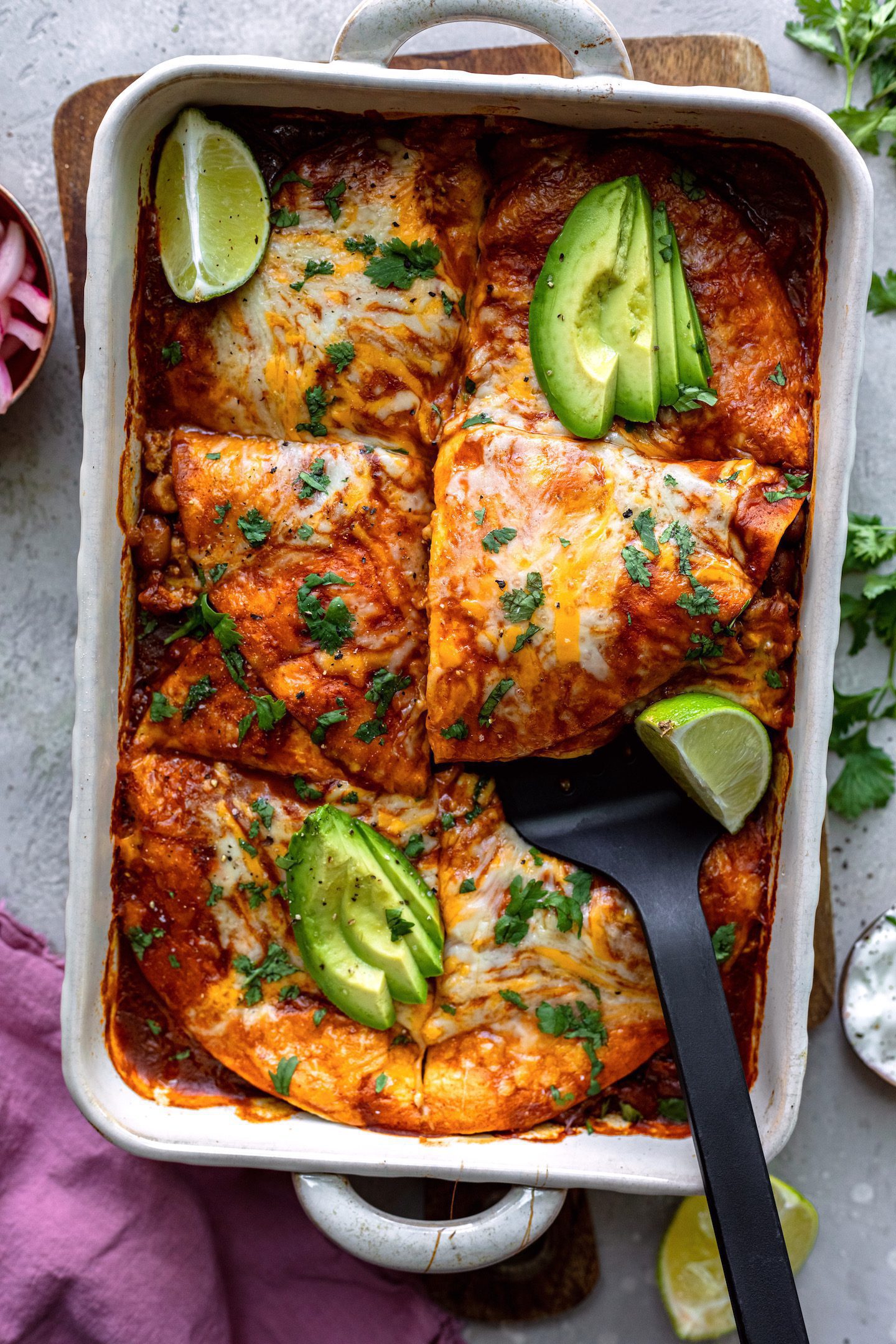 Overhead photo of an enchilada casserole cut into slices and garnished with lime wedges and sliced avocado.