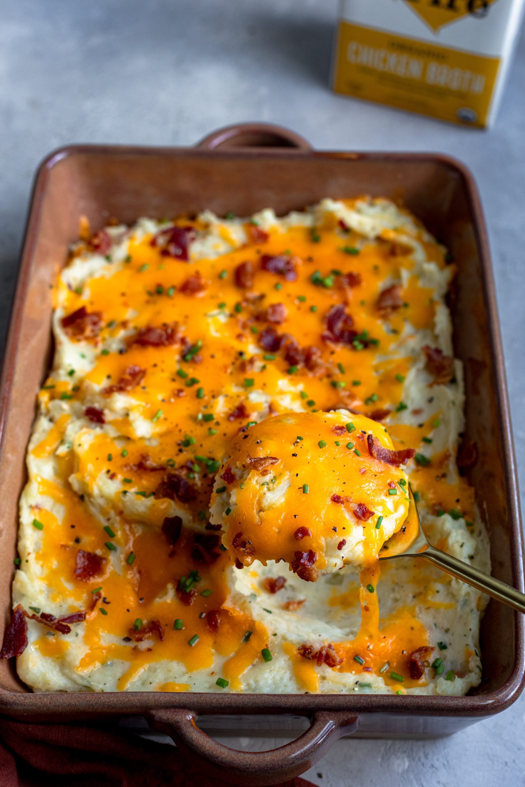 Close up of mashed potatoes topped with bacon and cheese being spooned out of a baking dish.