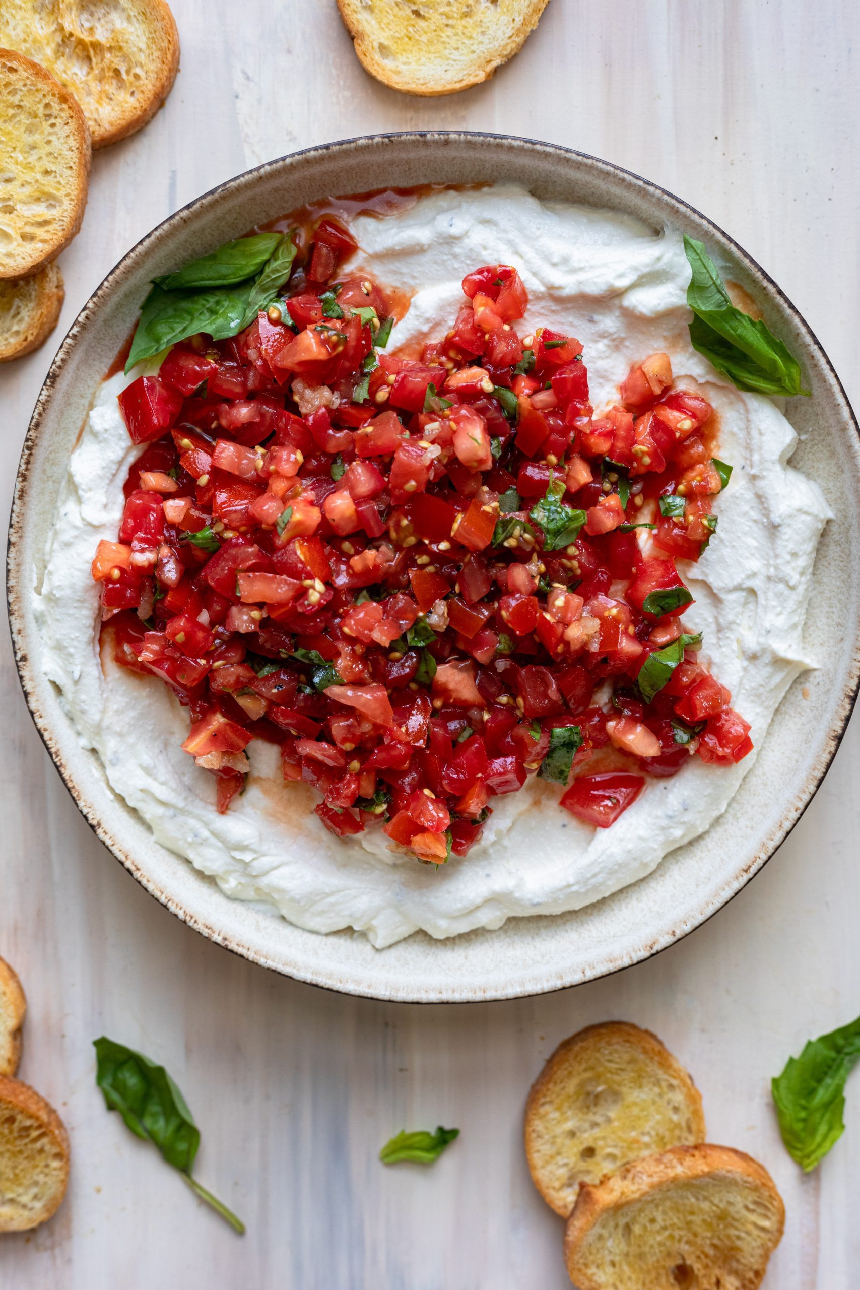 Bowl of Whipped Ricotta and bruschetta dip on a light wood background with basil leaves and crostini.