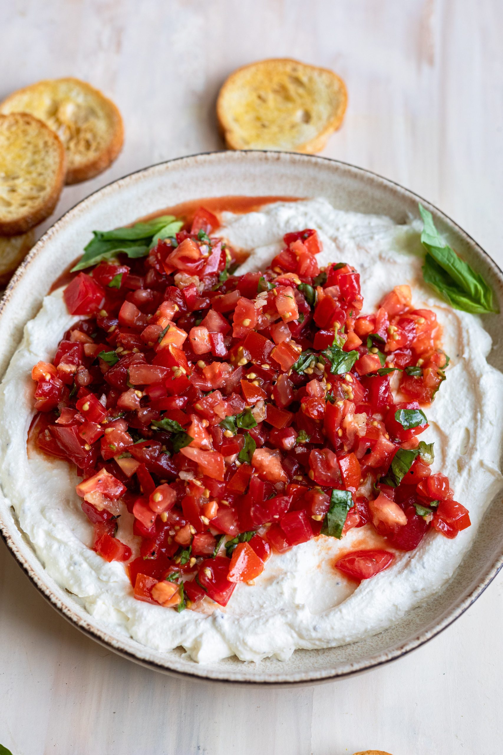 Whipped ricotta bruschetta dip with crostini on a light wood background.