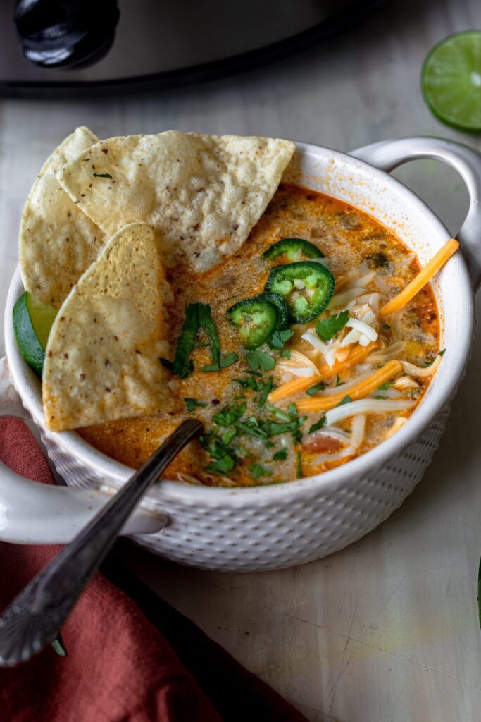 Bowl of White Chicken Chili garnished with cilantro, shredded cheese, jalapenos and tortilla chips, with a spoon to eat.