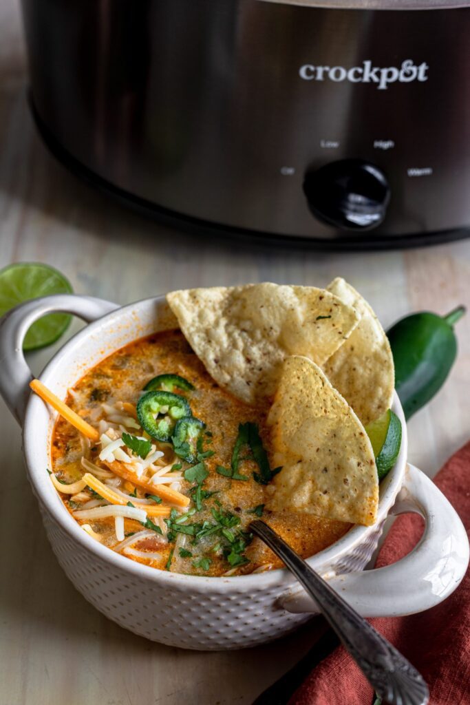 Bowl of white chicken chili topped with cheese, cilantro, jalapenos and tortilla chips with a CrockPot in the background.