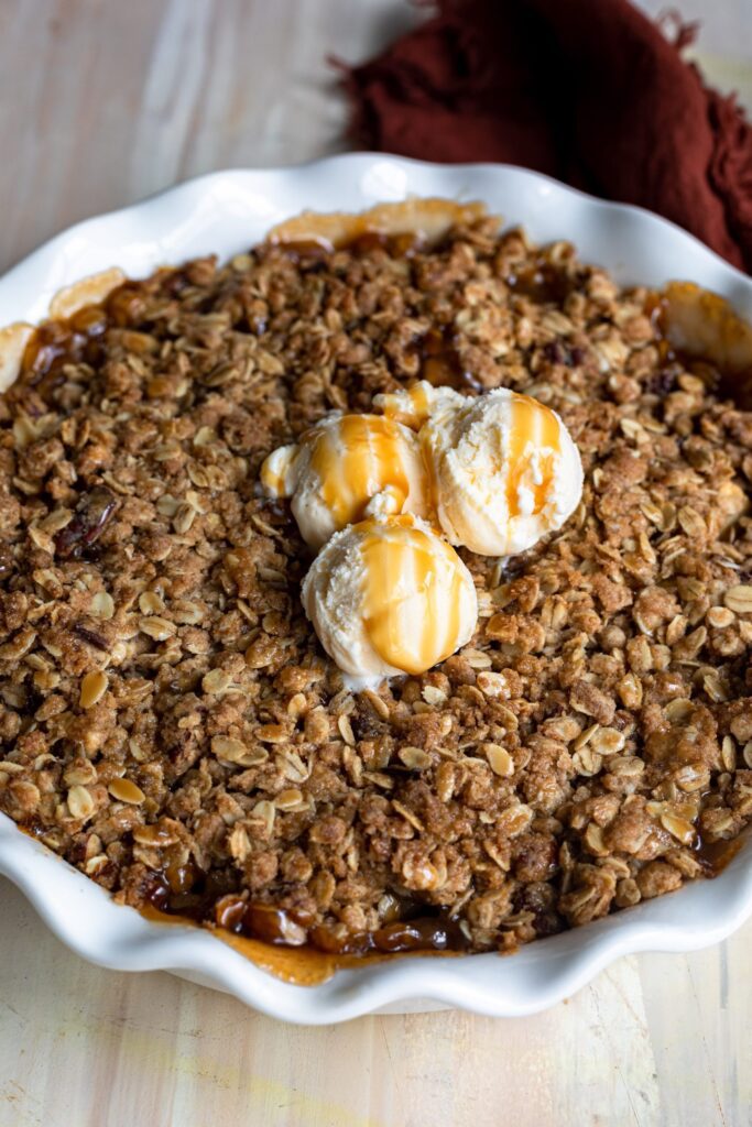 A white bowl filled with Cinnamon Apple Crisp with Pecans and Oats topped wih Caramel Drizzle and Vanilla Ice Cream.