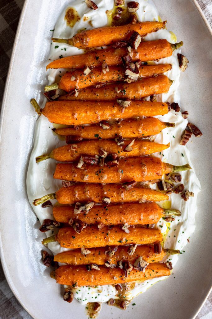 A baking dish filled with roasted carrots in a maple mustard glaze on a bed of whipped goat cheese, topped with chopped pecans.