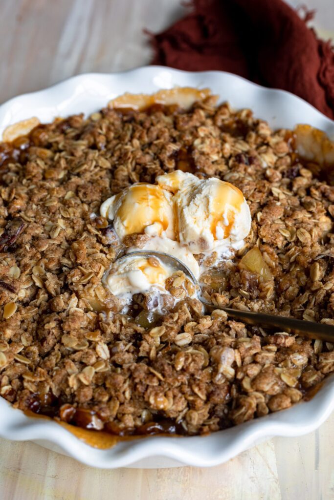 A bowl of oaty Pecan Bourbon Apple Crisp topped with Vanilla Ice Cream and smothered in Caramel Sauce.