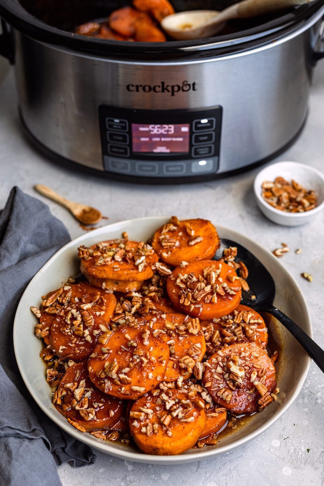 A plate of candied sweet potatoes topped with chopped pecans, showing a CrockPot slow cooker in the background. 