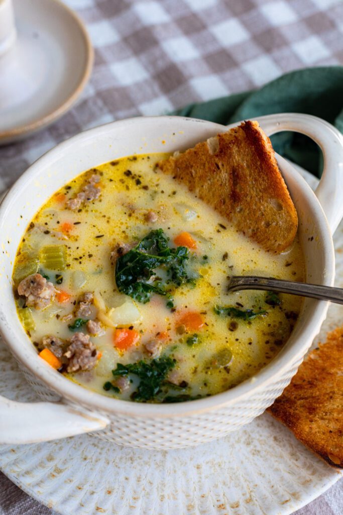 A bowl filled with colorful Sausage Tuscan Soup, topped with fresh Kale and toasted bread dipped in.