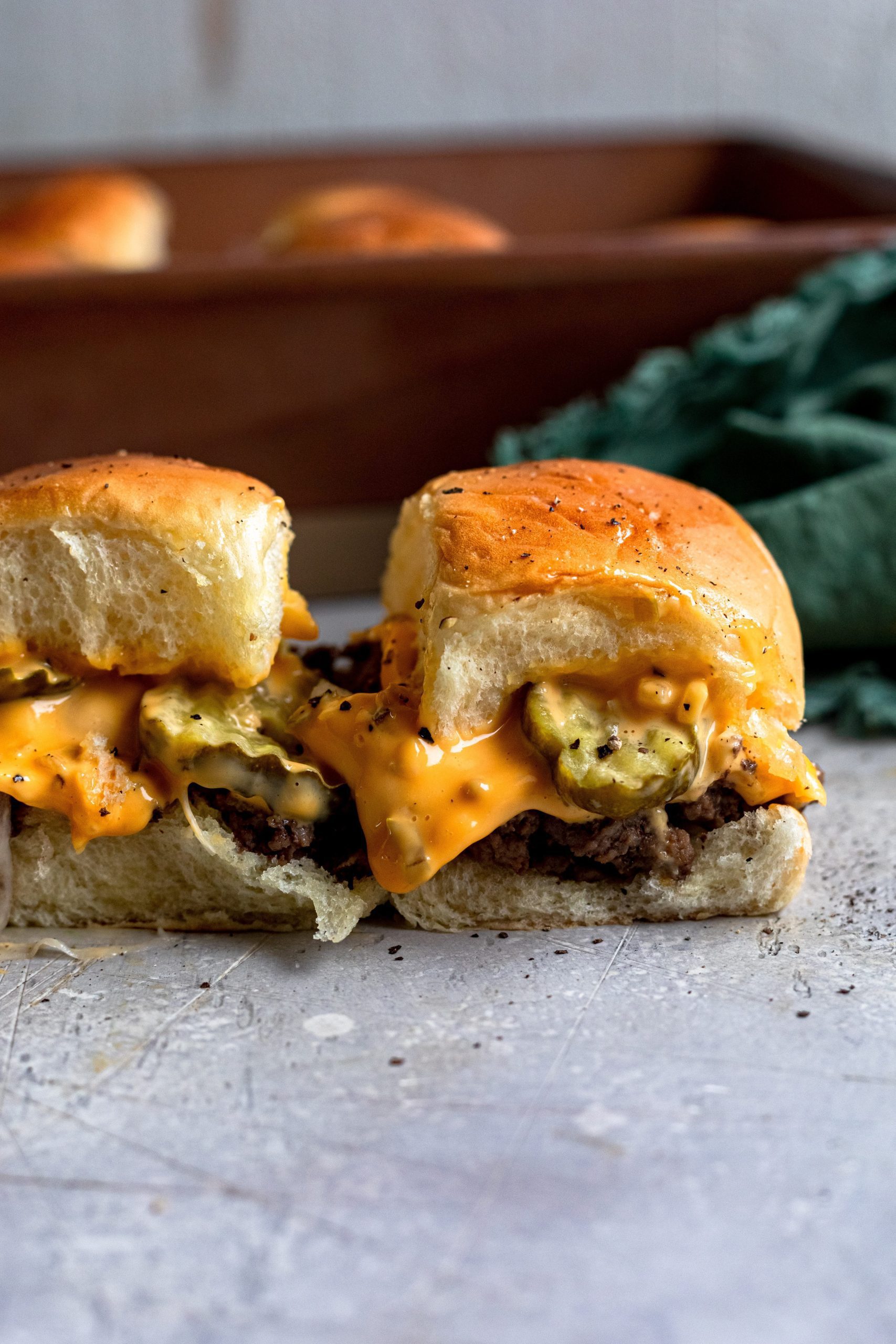 Side profile of 2 baked cheeseburger sliders showing glossy bun, melted cheese, pickle and juicy beef.