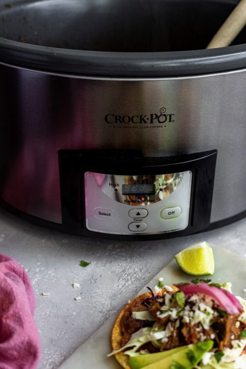 A large silver Crock-Pot with a freshly prepared barbacoa beef stacker nearby on a tray.