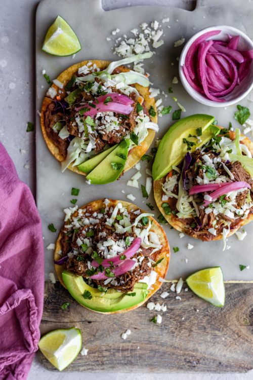 A trayful of colorful mini beef tacos topped with fresh cilantro, cotija cheese, pickled red onion, refried beans and avocado. 