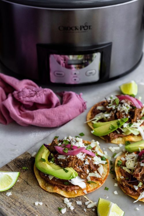 Freshly assembled barbacoa beef tostadas with a large Crock-Pot slow cooker in the background.