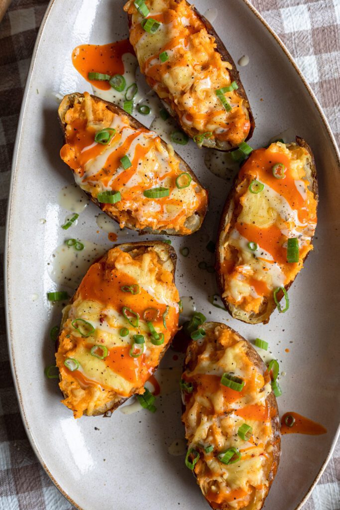 5 halved baked potatoes with buffalo chicken, melted cheese, ranch and spring onion garnish arranged on a white serving tray.