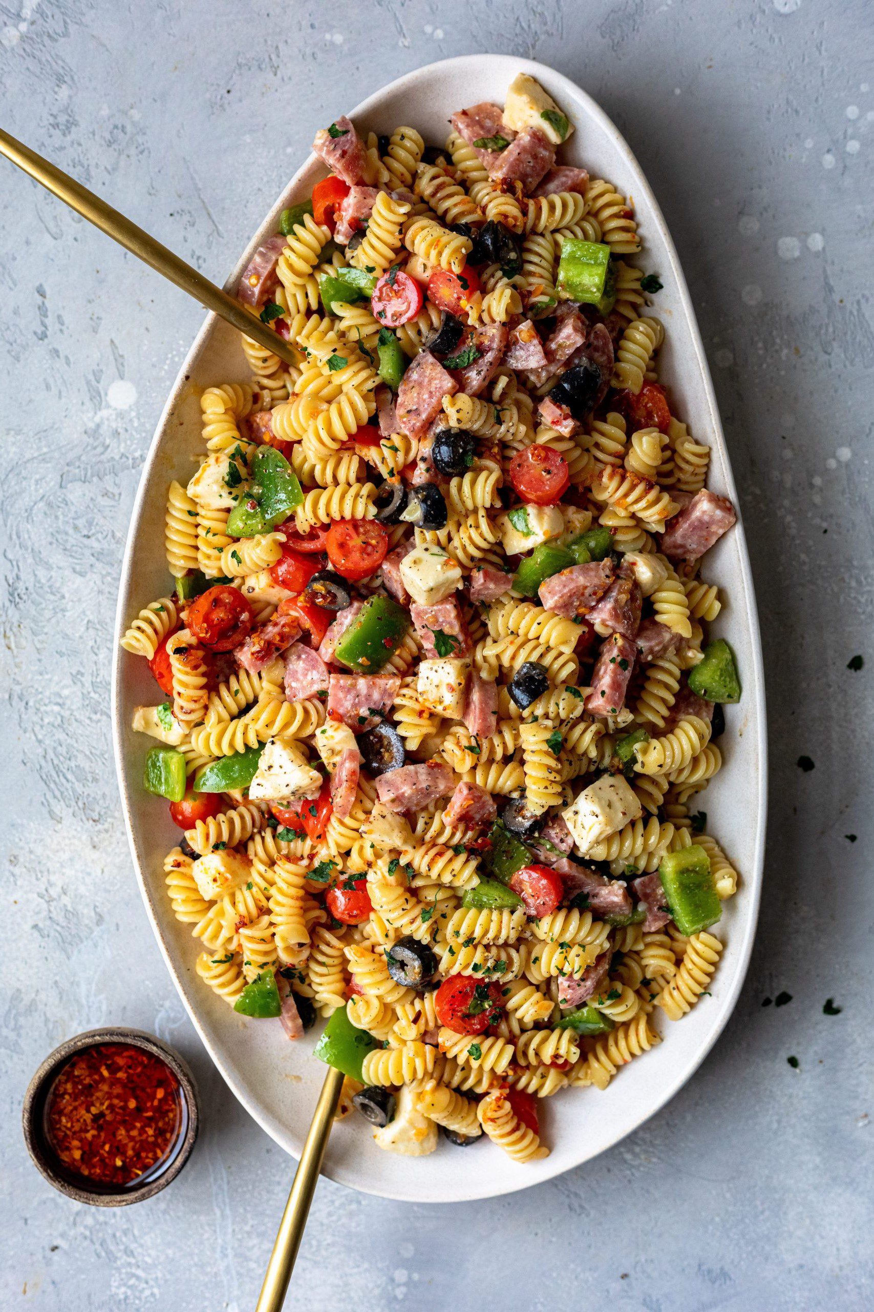 A serving platter full of pasta salad made with cooked italian meats, green bell peppers and sliced black olives.