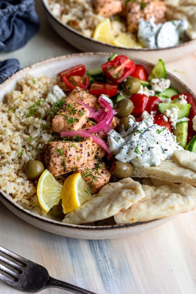 A shallow dish filled with a Greek inspired salmon and rice bowl, topped with vibrant fresh vegetables and tzatziki.